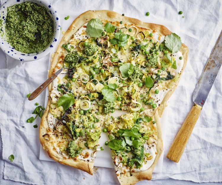 Op een witte snijplank ligt de rechthoekige flammkuchen. Hij heeft prachtige groene tinten vavn de erwtjes, prei en blaadjes waterkers. Er ligt een groot mes langs om aan te snijden een potje met extra pesto van waterkers.
