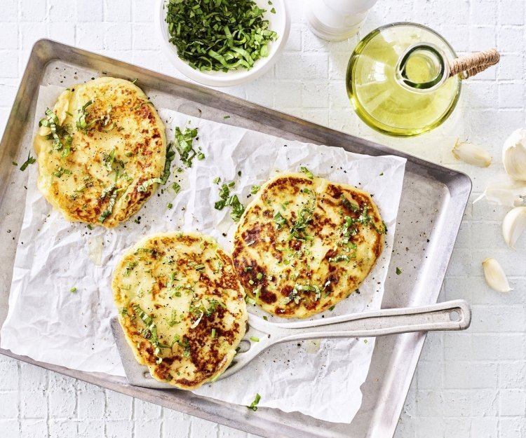 Drie broodjes zijn gebakken, en wat voor een! Flatbread gemaakt van een restje aardappelpuree liggen te blinken op een grijze bakplaat met wit boterpapiertje op. Verse kruiden liggen klaar om af te werken. 