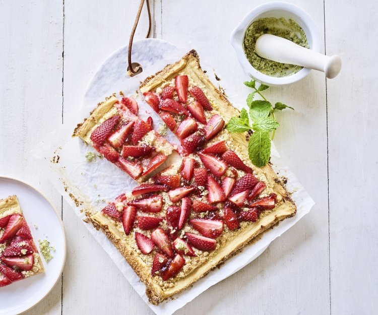 Frangipanetaart met aardbeien Een heerlijke plaattaart stat op tafel. Deze frangipanetaart ligt op een marmere snijplank en is afgewerkt met stukjes verse aardbeien en homemade muntsuiker. De taart is aangesneden en er ligt een stukje op een bord.