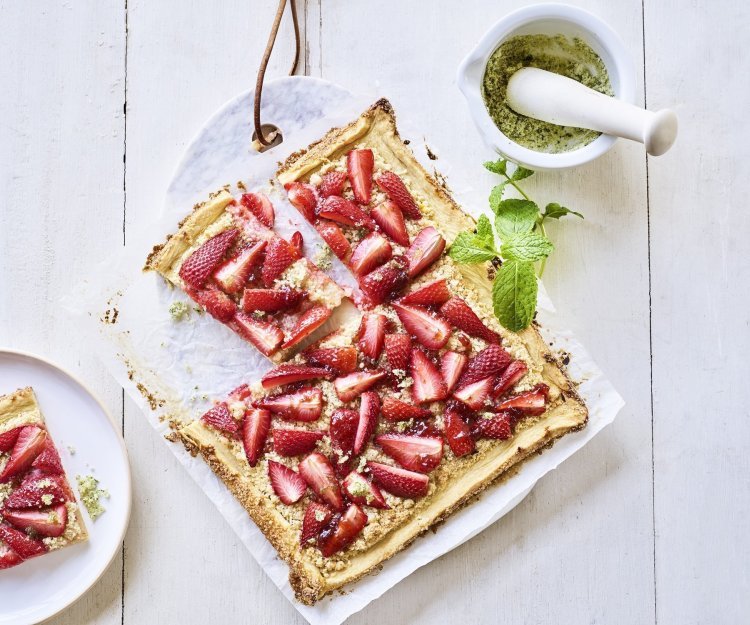 Een heerlijke plaattaart stat op tafel. Deze frangipanetaart ligt op een marmere snijplank en is afgewerkt met stukjes verse aardbeien en homemade muntsuiker. De taart is aangesneden en er ligt een stukje op een bord. 