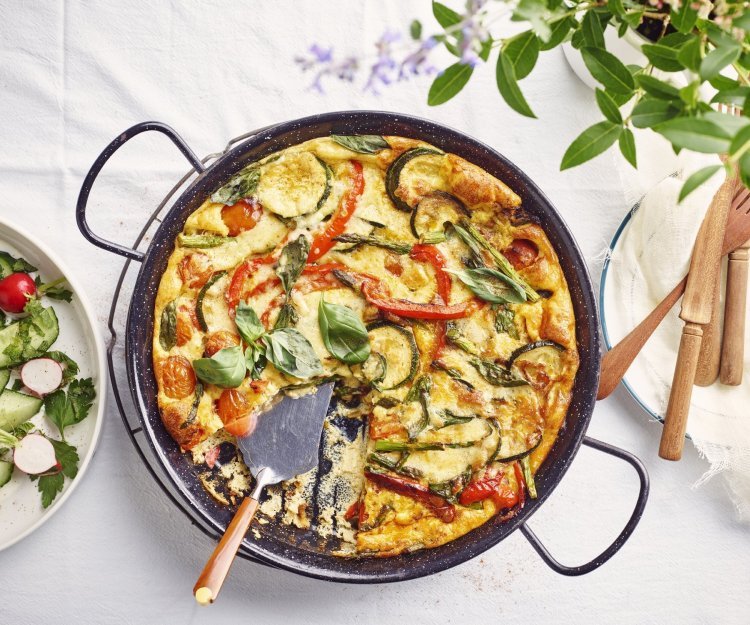 Frittata met kerstomaten, paprika, courgette en groene asperges De frittata staat op tafel in zwarte pan, waardoor hij nog mooier oogt dan dat hij al is. De tomaatjes, paprika, courgette en asperges geven leuke kleurtjes aan de frittata. Er staat een slaatje langs de linkse zijde, met komkommer, radijsjes en verse kruiden.