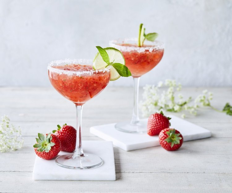 Cocktail time! Wij serveren een frozen daiquiri met verse aardbeien en basilicum. We presenteren hem in een wijnglaasje met suikerrandje, schijfje limoen en basilicumblaadje. Er liggen enkele aardbeien langs samen met witte bloemetjes. Schol! 