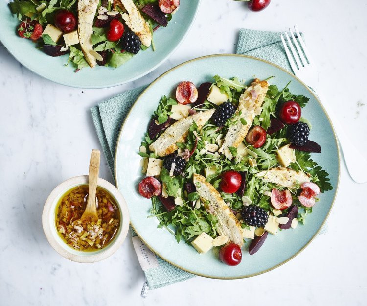 Een gemengde salade met sla, kersen, bramen en gekookte kipfilet gedresseerd op een lichtblauw bord. De dressing is er naast gezet in een keramieke potje met houten lepeltje in, de dressing bestaat uit sjalot, rode wijazijn, olijfolie en peper en zout. 