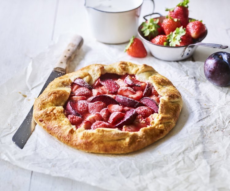 Galette met aardbeien en pruimen Een klaargemaakte galette staat op een houten witte tafel. Hij is prachtig goudgeel gebakken en het fruit oogt zoet-rode tinten. Er liggen nog wat aardbeien langs, alsook een paarse pruim.