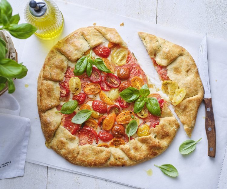 Galette met tomaten en mozzarella De galette van tomaten ligt op de tafel op een wit boterpapiertje. Hij is mooi gebakken en er is een stukje van gesneden. Er staat een basilicumplantje langs, alsook een flesje gouden olijfolie.