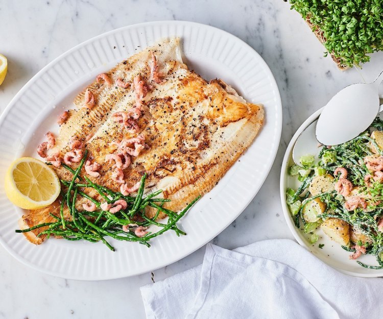 Gebakken schartong met zilte aardappelsalade Een heerlijk krokant visje afgewerkt met zeekraal ligt op een groot bord. Er zijn enkele garnaaltjes aan toegevoegd die mooi roze blinken. De aardappelsalade staat er langs in een witte kom met witte lepel. Om af te werken ligt er 1 citroen doormidden gesneden.