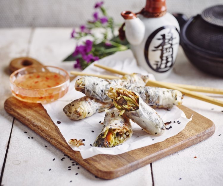 Gebakken springrolls met buikspek uit de airfryer Heerlijke krokante springrolls liggen op een houten snijplank. Ze zijn vergezeld met een zoetpikante dipsaus. In de achtergrond staat nog een theepot en paarse bloemetjes.