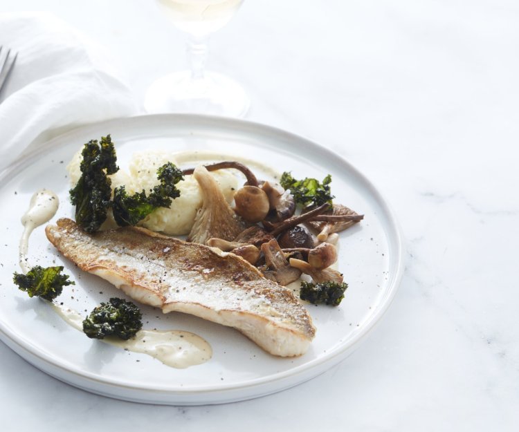 Gebakken wijting met knolselderpuree, boschampignons en krokante boerenkool Een gebakken visje op vel ligt op een wit bordje. Hij is afgewerkt met gebakken boschampignons en krokante stukjes boerenkool. Wij serveren hem met knolselderpuree en een glaasje witte wijn.