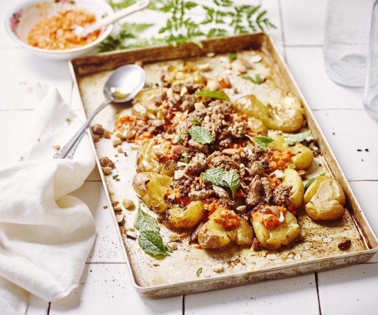 Op een ovenschaal liggen krokante crushed potatoes met homemade romesco saus bovenop en afgewerkt met lamsgehakt. De overige romesco saus staat bovenaan voor wie wat extra wil. Er ligt een witte servette op het aanrecht van witte Marokaanse tegeltjes. 