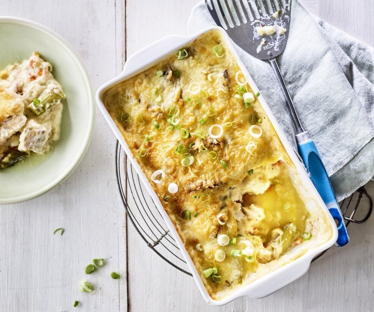 Gegratineerd vispannetje met Brugge kaas, asperges en venkel Het vispannetje staat op een metalen onderlegger op een witte houte tafel. De schepper ligt naast de schotel waaruit al 1 portie is uitgeschept in een lichtgroen bordje.