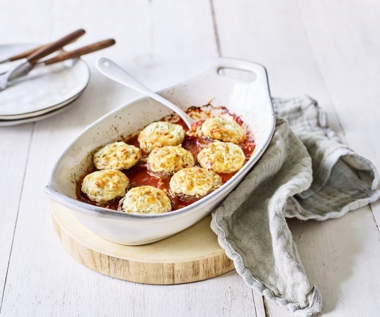 Gegratineerde champignons met blauwe kaas en tomatensaus Witte ovenschotel staat op een houten snijplankje op tafel. De ovenschotel is gevuld met de rode tomatensaus waarin de champignons mooi in geschikt zijn. Achteraan de foto zie je bordjes staan met bestek om aan de slag te gaan.