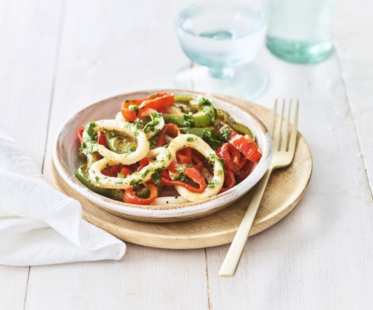 Gegrilde inktvis en paprika met kruidenolie Op tafel staat een keramiek bordje op een houten plankje, het bord is kleurrijk gevuld met groene- en rode paprikaringen, én niet te vergeten de inktvisringen. Een bordje vakantie, als je het mij vraagt.