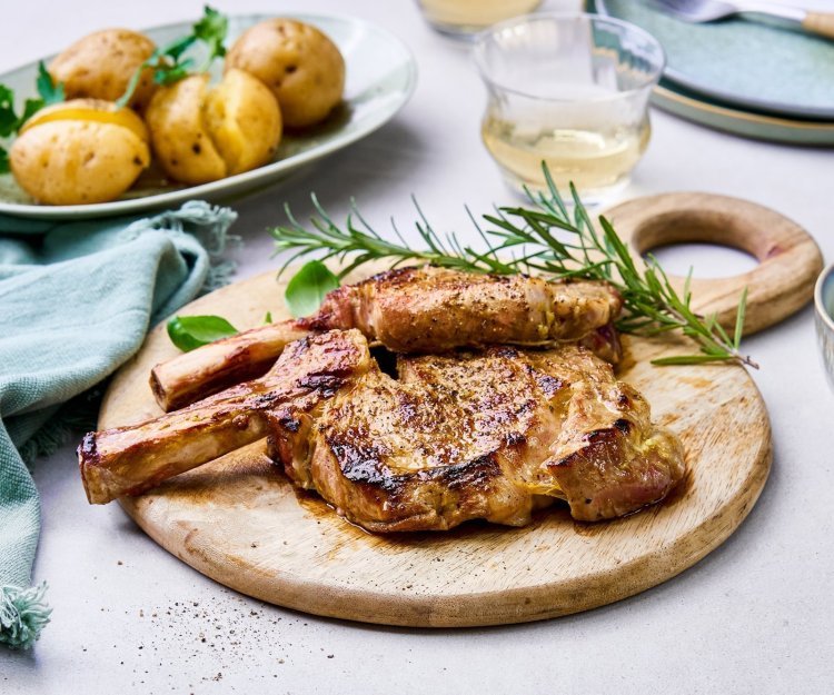 Gegrilde kalfskoteletten met salsa verde op de barbecue Gegrilde koteletten liggen op een houten ronde snijplank te blinken. Er ligt een takje rozemarijn langs, een bordje met de gegrilde krieltjes en een potje met de salsa verde om toevoegen aan het vlees.