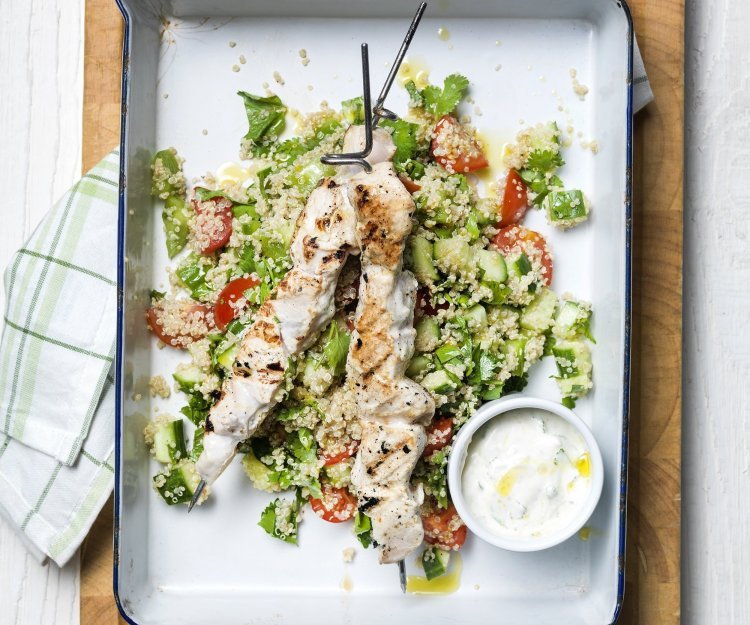 Gegrilde kippenspiesjes in yoghurt met quinoa-salade Grote witte bakplaat staat op tafel met eerst een quinoaslaatje en groentjes gepresenteerd. Hierop liggen de gegrilde brochettes van kip met een dressing van yoghurt.