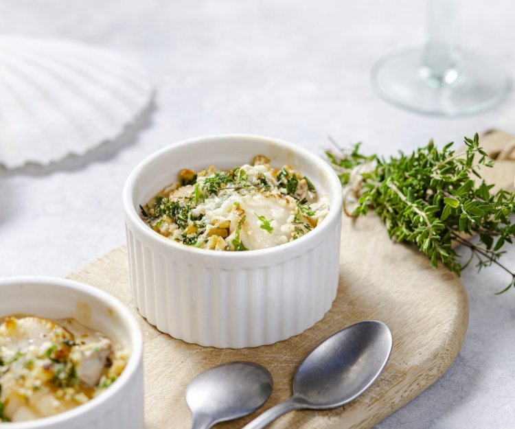 Gegrilde sint-jakobsvruchten met een crumble van crackers, kaas en verse kruiden Gegrilde sint-jakobsvruchten gepresenteerd in kleine witte ovenschaaltjes met een crumble van crackers, kaas en verse kruiden. Ze staan op een houten plankje met kleine lepeltjes en verse tijm naast.