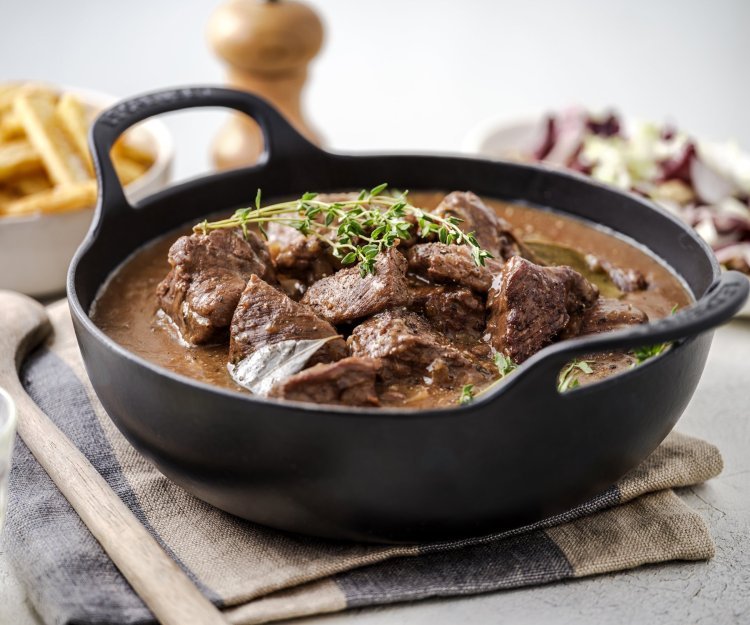 Gentse stoverij Er staat een zwarte sudderpot op tafel met Gentse stoverij, in de achtergrond zijn er frietjes te zien en een witloofslaatje.