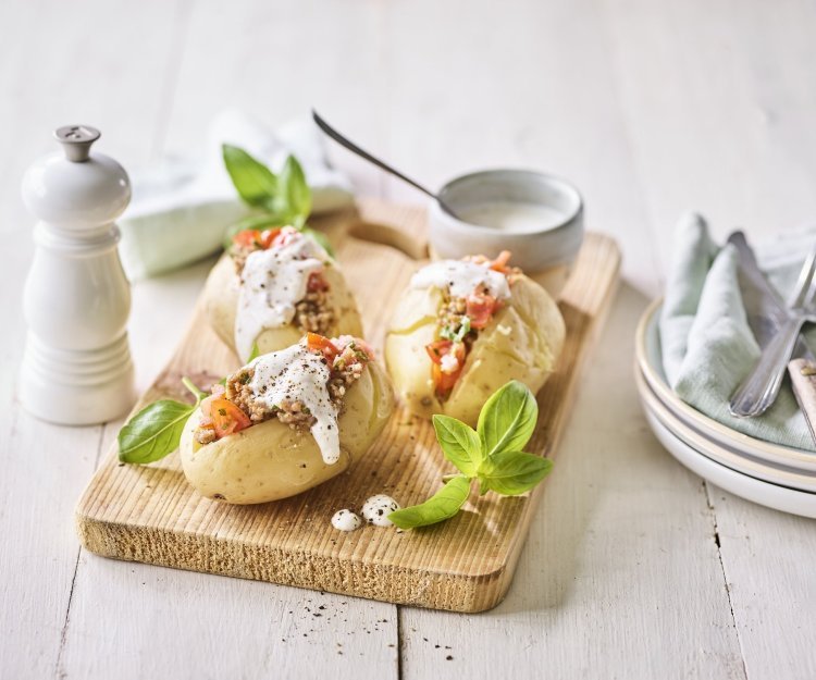 Enkele klaargemaakte en afgewerkte gepofte aardappelen liggen op een houten snijplank. Ze zijn gevuld met gehakt, stukjes tomaat en afgewerkt met een geitenkaasdip. Naast de aardappelen staan enkele bordjes, bestek en servetten om meteen te beginnen. 