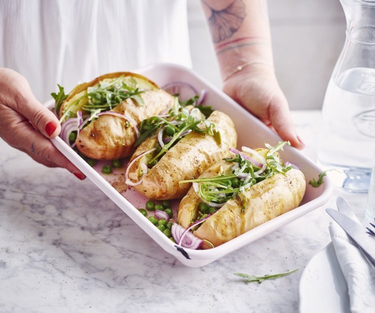 3 gepofte aardappelen met pesto, erwtjes en rucola liggen in een witte ovenschaal. De vrouw wil de ovenschaal plaatsen op tafel.