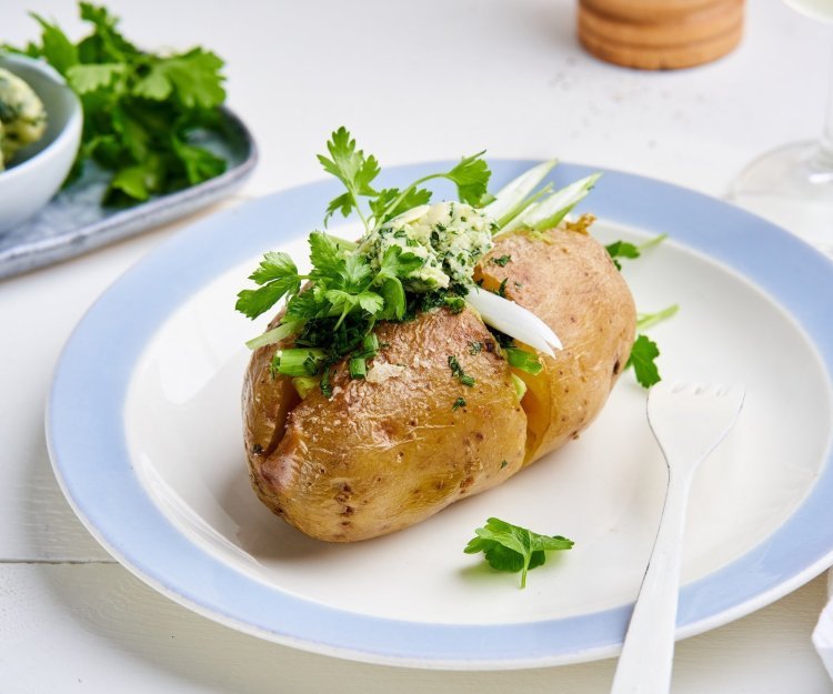 Gepofte aardappelen met kruidenboter Een gepofte aardappel ligt op een bordje, gevuld met homemade kruidenboter, lente-ui en verse kruiden.