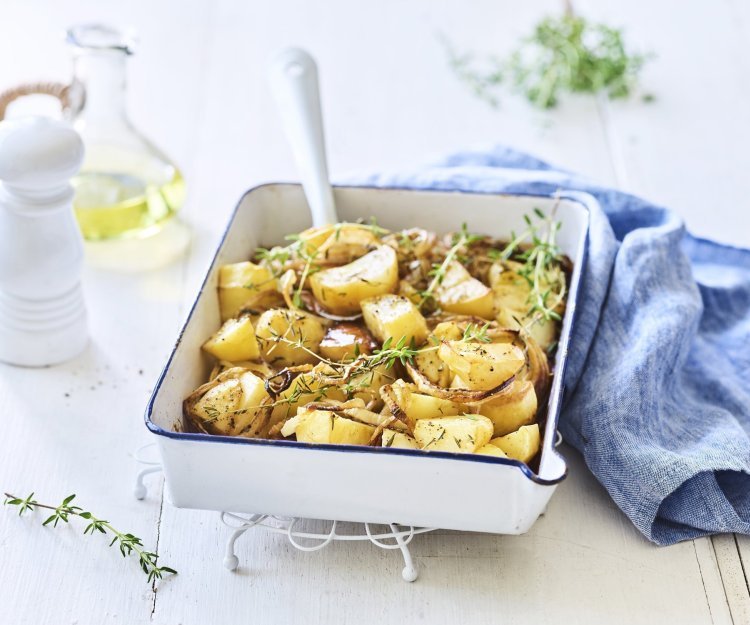 Een witte ovenschaal met donkerblauw randje staat op een keukenonderlegger op een witte houten tafel. Er staat een witte pepermolen en een glazen flesje met olijfolie langs. De verse tijm zorgt voor extra kleur bij de geroosterde aardappelen, er ligt ook nog een blauwe linnen servette langs de ovenschotel.