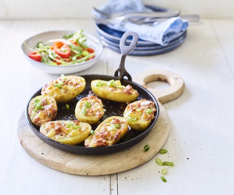 Gevulde aardappelen met spek en Flandrien kaas op de barbecue 7 gevulde aardappelen liggen op een zwarte vuurvaste pan, ze zijn gevuld met aardappelkruim, kaas en spekblokjes. Er ligt een handdoek achteraan in beeld en een slaatje met tomaatjes.
