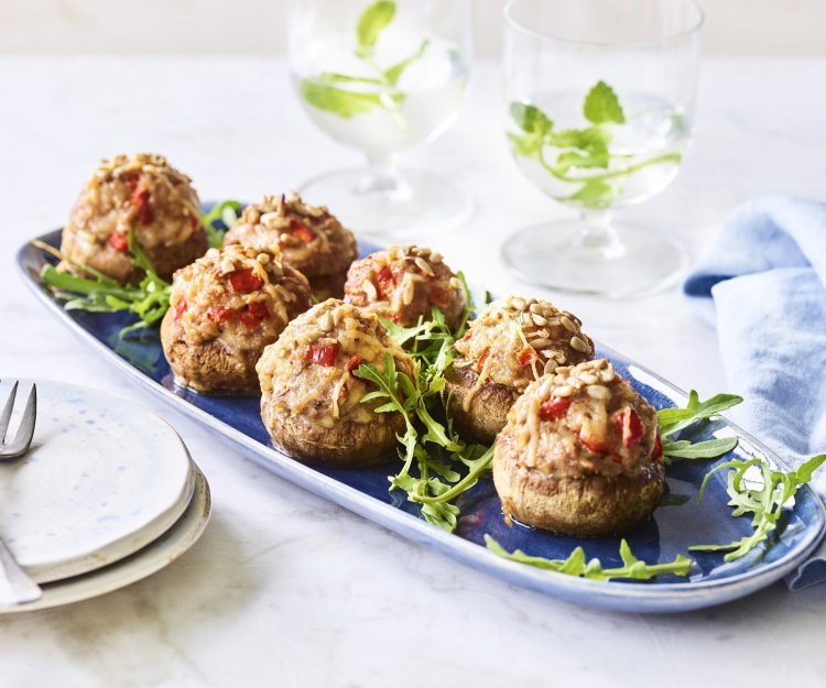 Gevulde champignons met gehakt, paprika en zonnebloempitten Op een blauw schaaltje liggen gevulde champignons. Ze zijn geserveerd met enkele blaadjes rucola en een stappeltje bordjes met mini-vorkjes om het hapje proper te eten.