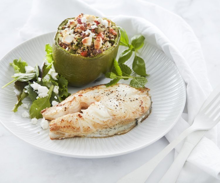 Een gebakken moot van schelvis ligt op een wit bord. Hij is geserveerd met een gevuld groene paprika. Deze is opgevuld met quinoa, tomaten- en komkommerstukjes en geitenkaasblokjes.