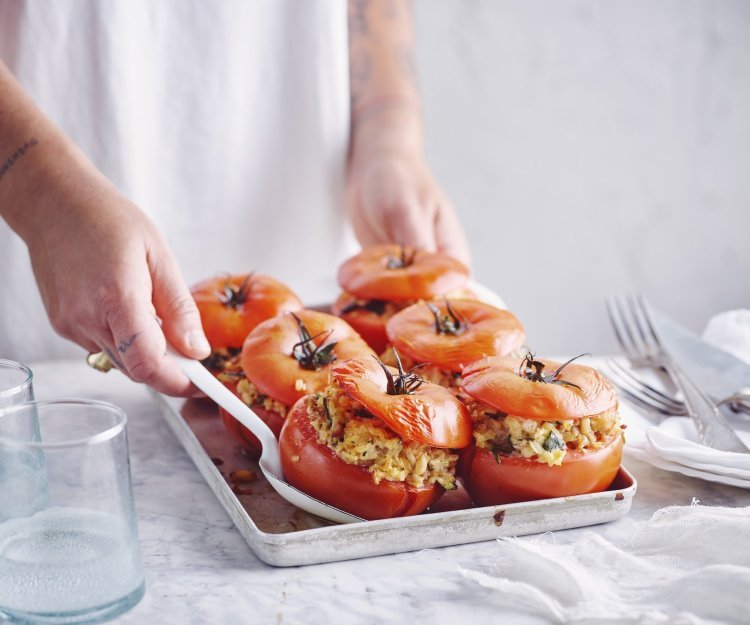 Gevulde tomaat met rijst, spinazie, courgette en platte kaas Zes gevulde rode tomaten in een ovenschaal zijn aan tafel gebracht door een vrouw in witte t-shirt. Ze schept alvast eentje op voor zichzelf.