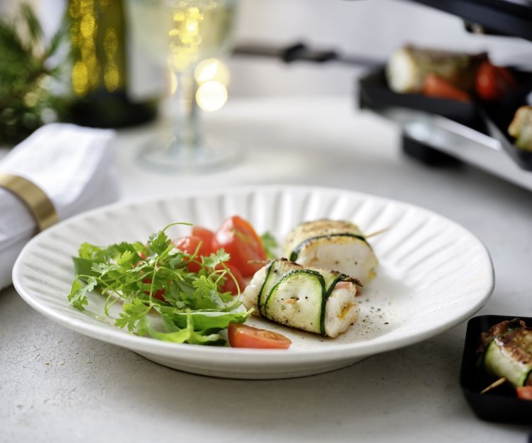 Gourmet van wijtingpakketje met gepofte kerstomaatjes Twee pakjes liggen op een wit bord. Het pakje is gemaakt van een stukje vis verpakt in een sliertje courgette. We serveren dit feestelijk gerechtje met een frisgroene kruidensla en kertomaatjes.