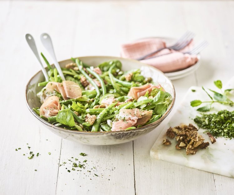Een licht groene serveerkom is gevuld met onze groene salade boordevol, jawel, groene groenten. Denk maar aan verse sla, boontjes, paprika en erwten. De salade is afgewerkt met rozige gerookte hamsneetjes en verse groene kruiden.  