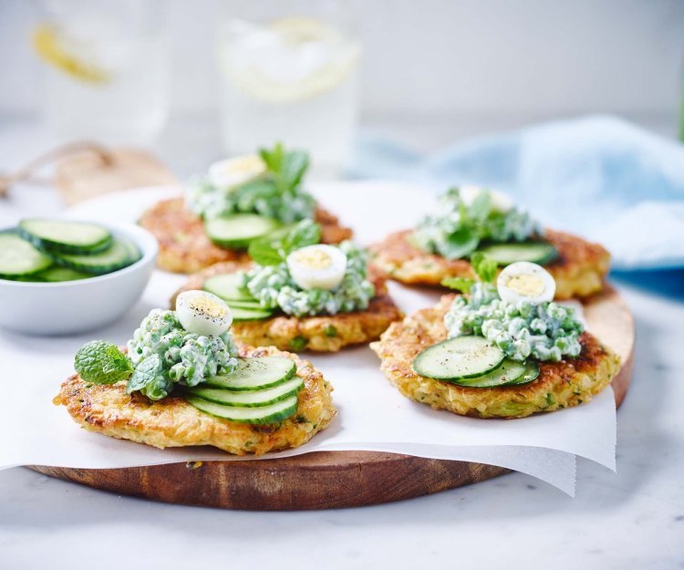 Groentekoekjes van pastinaak, courgette en erwtjes op een houten plank gepresenteerd. Er ligt een licht blauwe linnen servette langs en een waterkaraf met glazen.