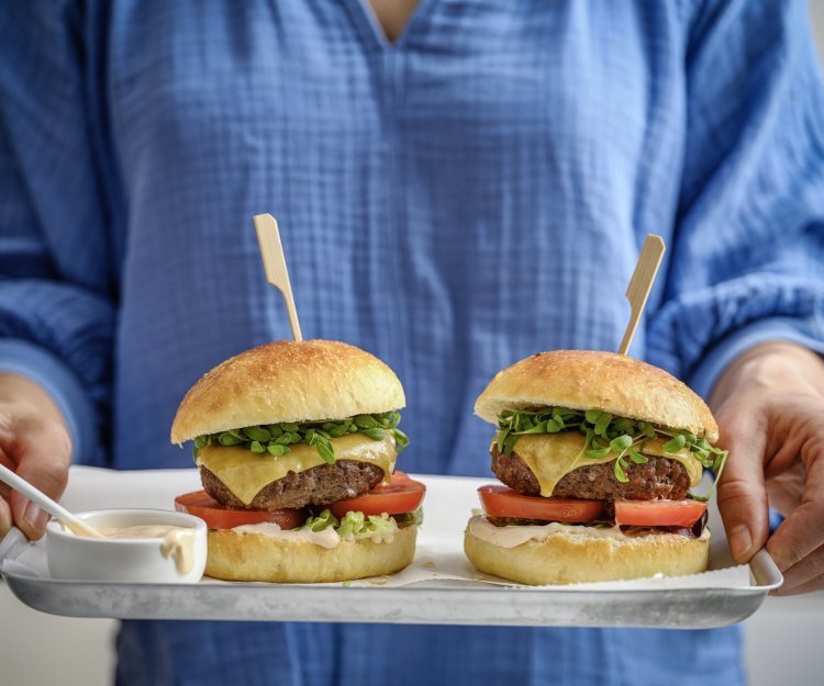 Hamburger Potato buns Een vrouw staat in beeld met haar romp draagt een plateau met hierop twee hamburgers. De hamburgerbroodjes hebben een mooi goudgele kleur, ze werden gemaakt met aardappelpuree in het deeg. Verder zijn de burger belgd met een schijfje vlees, kaas, tomaat, sla en saus natuurlijk.