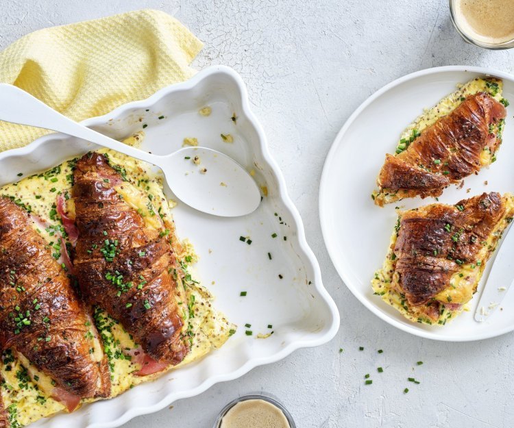 Hartige croissants uit de oven met ham, kaas en mosterd Er staat een witte ovenschotel op tafel gevuld met croissants. Ze zijn doormidden gesneden, bestrijkt met mosterd en belegd met ham en kaas. Daarna is een beslag toegevoegd en werden ze 10 minuten in de oven geplaatst. Om vingers en duimen van af te likken, al zeg ik het zelf.