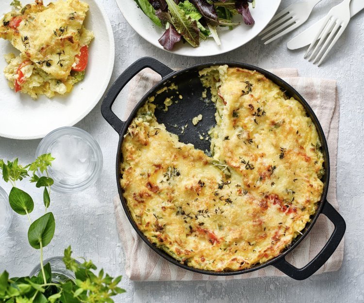 Een taart van aardappelpuree en paprika, staat op het midden van de tafel. Hij zit in een zwarte schotel en heeft een mooi kaaskorstje meegekregen uit de oven. Er is al 1 portie uitgeschept, en er ligt ook bestek op tafel, sla en verse kruiden. 