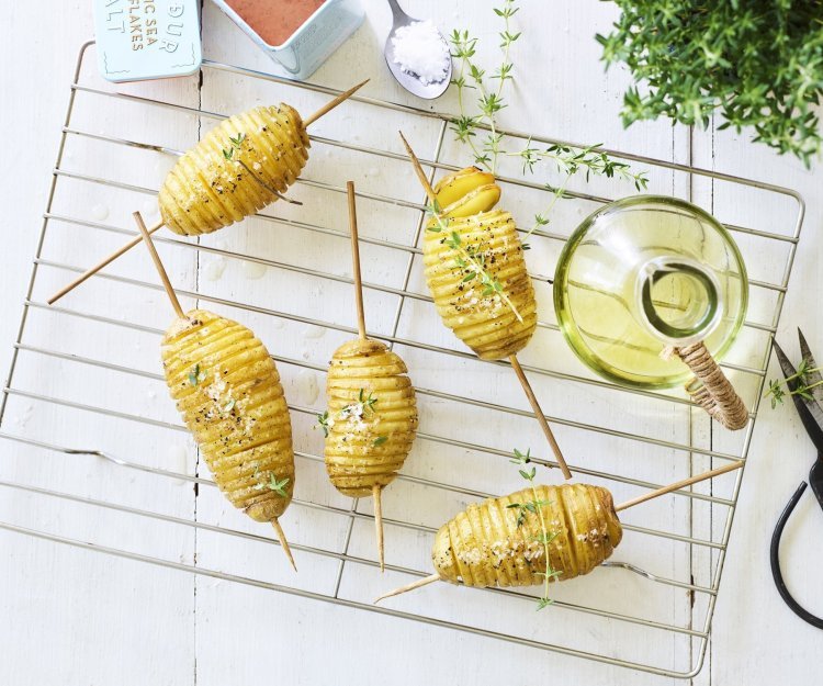 Hasselback potatoes liggen op een bakrooster te blinken. Ze zijn doorprikt met lange houten prikker om makkelijker te kunnen draaien op de BBQ. Er staat een flesje olie, verse tijm en een potje zeezout naast. 