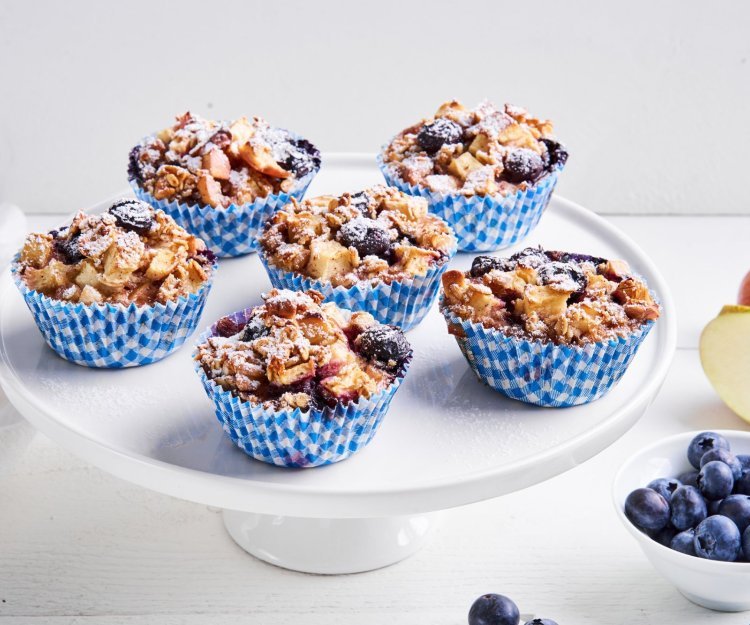 Op een taartenserveerschaal staan 6 muffins in blauw-witte muffinpapiertjes. Er ligt nog een appel langs en blauwe bessen in een potje. 