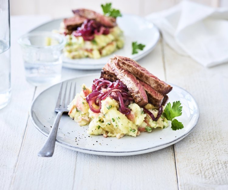 Hete bliksem met biefstuk en rode ui Een lichtblauw bordje is gevuld met een combinatie van puree en appelmoes. Deze puree is afgewerkt met gebakken rode ui en stukjes vlees. Er staan 2 van deze bordjes op tafel met een glas water.