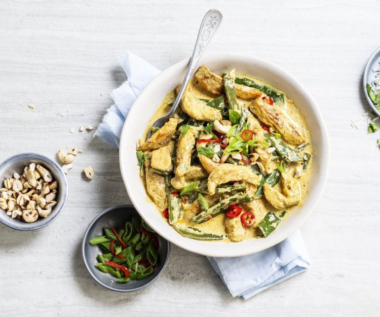 Kip curry met peultjes en noten Er staat een grote witte serveerschaal op tafel gevuld met de goudgele kip curry. Je ziet de groene peultjes en rode chili duidelijk. Langs het gerecht staan nog extra noten, gesneden lente-uitjes en een bordje rijst.