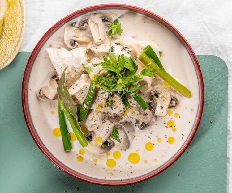 Kip in melk met champignons en ui Kip gegaard in melk staat op tafel in een diep beige bord met bordeau randje. Er liggen lente-uitjes en champignons bij. Het bordje staat op een blauwgroene onderlegger.