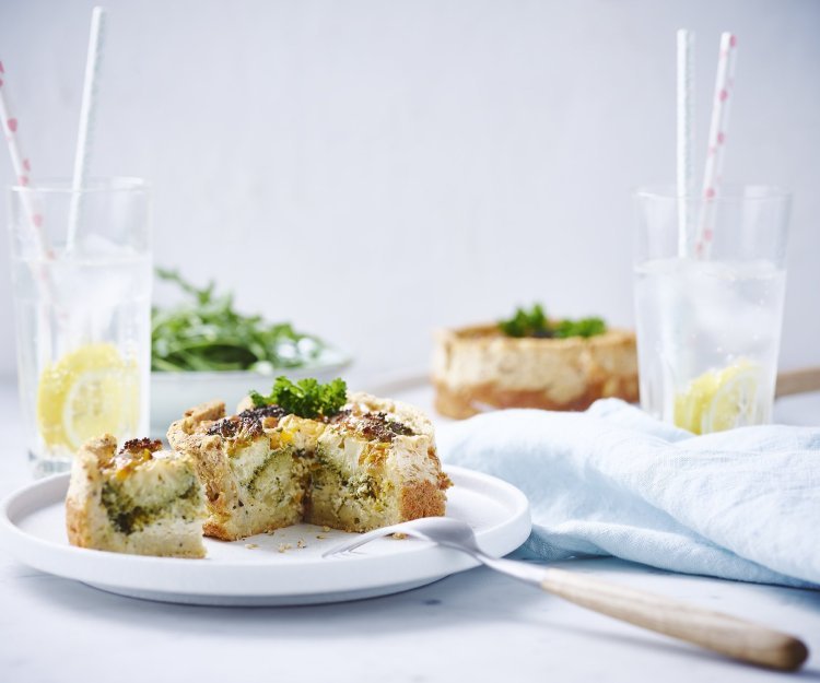 Kleine quiche van brood met broccoli en paprika op wit bordje met houte bestek langs het bord. Er staan ook twee waterglazen met een schijfje citroen en kartonnen rietje in.