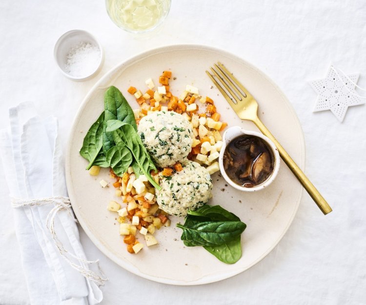 Knoedels, gemaakt van brooddeeg, liggen bovenop gestoofde blokjes peterseliewortel en zoete aardappel. Deze vegetarische ballen worden opgediend met een sausje van bospaddenstoelen. Het sausje staat mee op het bord in een potje. Goud bestek en kerststerren liggen langs het bord.