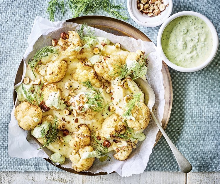Knolselderpuree met geroosterde bloemkool, groene kruidendressing en noten Puree van knolselder is uitgeschept op een groot lichtbruin bord. Hierop zijn de geroosterde bloemkoolroosjes geserveerd met wat drizzle van de groene kruidendressing, noten en verse dille. De overige kruidendressing is in een witte potje en staat langs het bruine bord, samen met nog wat gehakte noten in een ander potje. Alles staat op een een plaid in een blauwgrijze kleur.