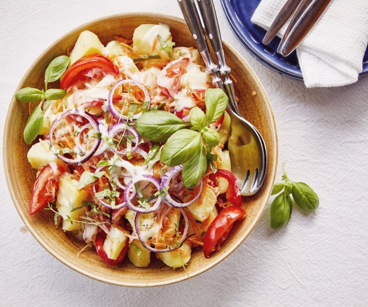 Koude aardappelsalade met tomaat en paprika Een grote serveerschaal staat op de tafel, ze is gevuld met gekookte aardappelstukjes, tomaat, paparika, rode ui en wortel. De dressing is al toegevoegd en verse kruiden zoals basilicum en tijm geven een nog extra kleurtje aan de salade.