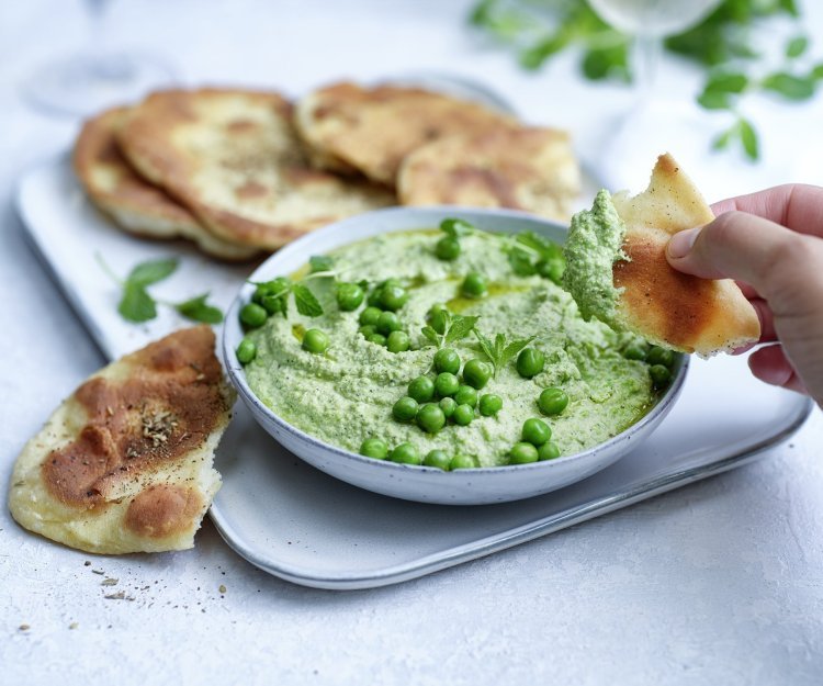 Krokant platbrood met erwtendip Krokant gebakken platbrood ligt op een serveerschaaltje waarbij een dip van erwten en roomkaas in geserveerd. De dip is gemixt en heeft een mooie groene kleur, om af te werken zijn er nog enkele verse erwtjes bovenop gestrooid. Een hand neem alvast een stukje platbrood om te dippen in gezonde groentedip.