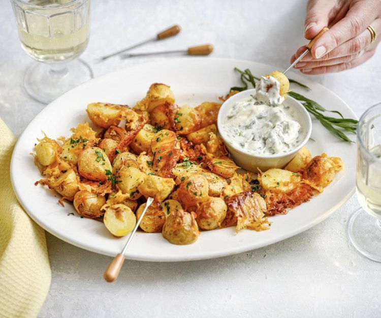 Krokante aardappelen in de oven met kaas en dragonmayonaise Een groot wit bord staat midden op de tafel, het is gevuld met heerlijke krokante aardappeltjes met kaas en een dipsausje van dragonmayonaise. Er is een hand die er eentje prikt en er staan twee glaasjes wijn. De aperitief is gestart.