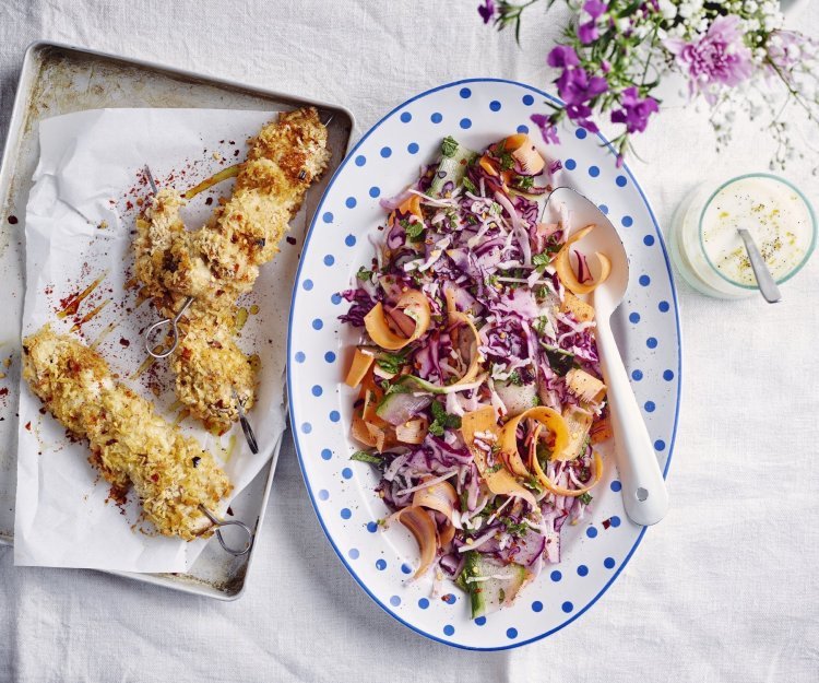 Krokante kipbrochette met een slaatje van kool, komkommer en wortel Krokante kipbrochette ligt nog de sidderen op een ovenplaatje. Daarnaast staat een kleurrijk groot bord met een salade op basis van rode kool, wortel en komkommer. De dressing op basis van yoghurt staat langs de salade.