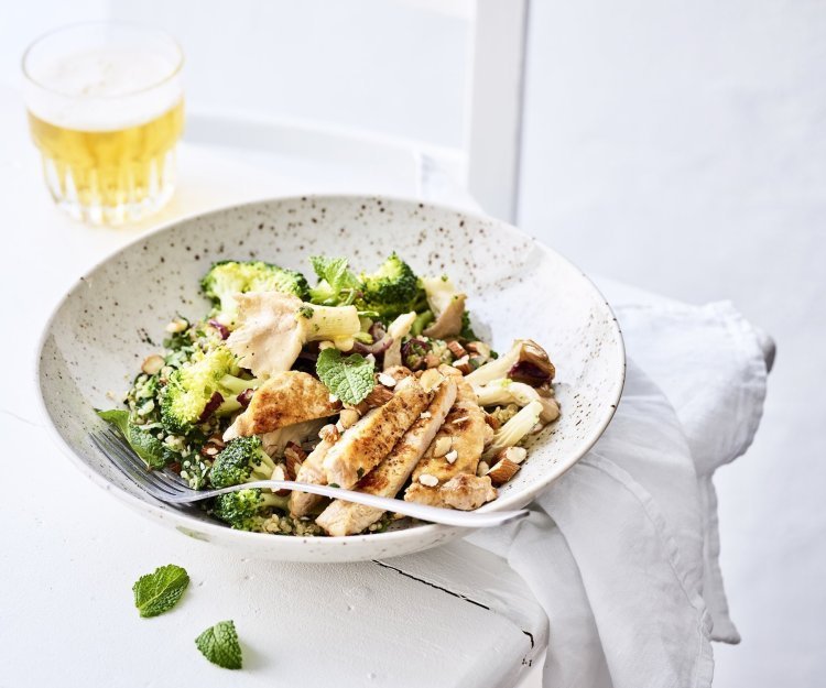 Lauwwarme quinoasalade met groenten, verse kruiden en varkensvlees Een rijkgevulde quinoasalade boordevol verse groenten en kruiden zoals broccoli, paddenstoelen, rode ui, peterselie, koriander en munt. Bovenop de quinoa werd het varkensvlees geplaatst in schijfjes. Er staat een witbier bovenaan het bord, op de tafel.