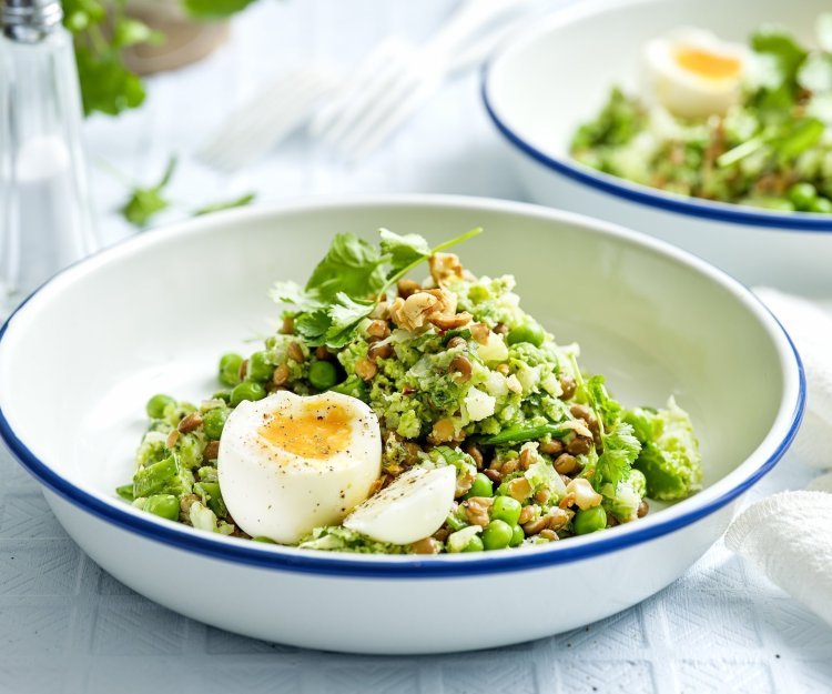 Een wit emaille bordje met blauwe rand is gevuld met de linzensalade. Het is een salade rijk aan groene groenten. Het gerecht is afgewerkt met een gekookt eitje. 