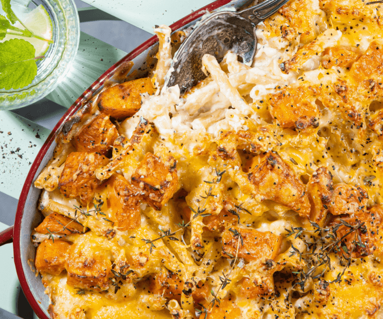 Mac en cheese met zoete aardappel Een grote bordeaukleurige ovenschotel staat op een licht blauwe stoel. De ovenschotel met ma&cheese en zoete aardappel is oranje kleurig. Boven de ovenschotel staan twee glaasjes water met ijsblokjes en verse munt.