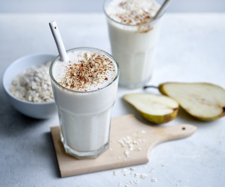 Twee klaargemaakte milkshakes staan op het lichtgrijze aanrecht te pronken. Er ligt een peer langs die doormidden gesneden is en wat havermoutvlokken gemorst. De shakes zijn afgewerkt met wat kaneelpoeder. 